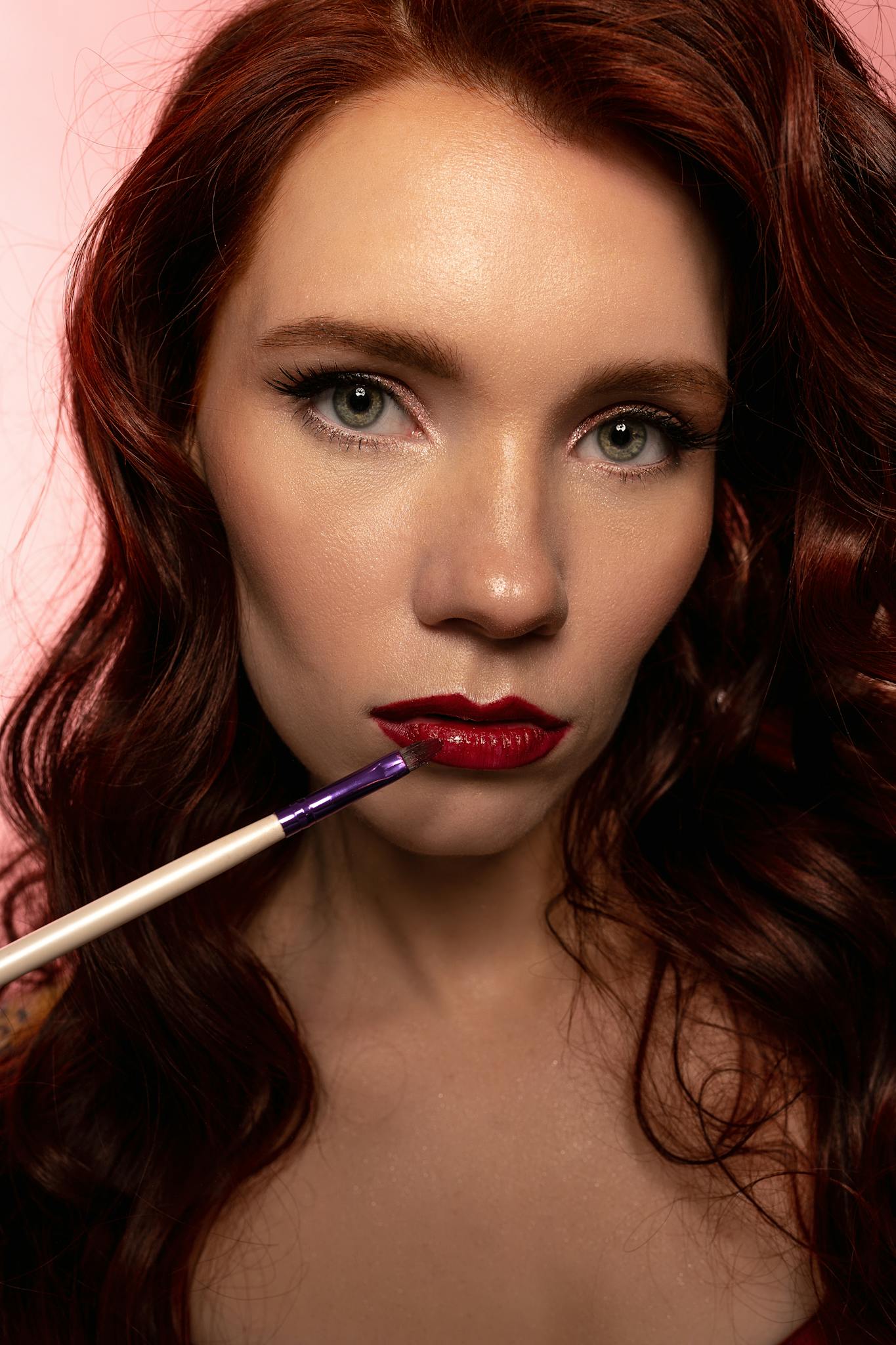 Close-up portrait of a beautiful redhead woman applying red lipstick, highlighting her flawless makeup and features.